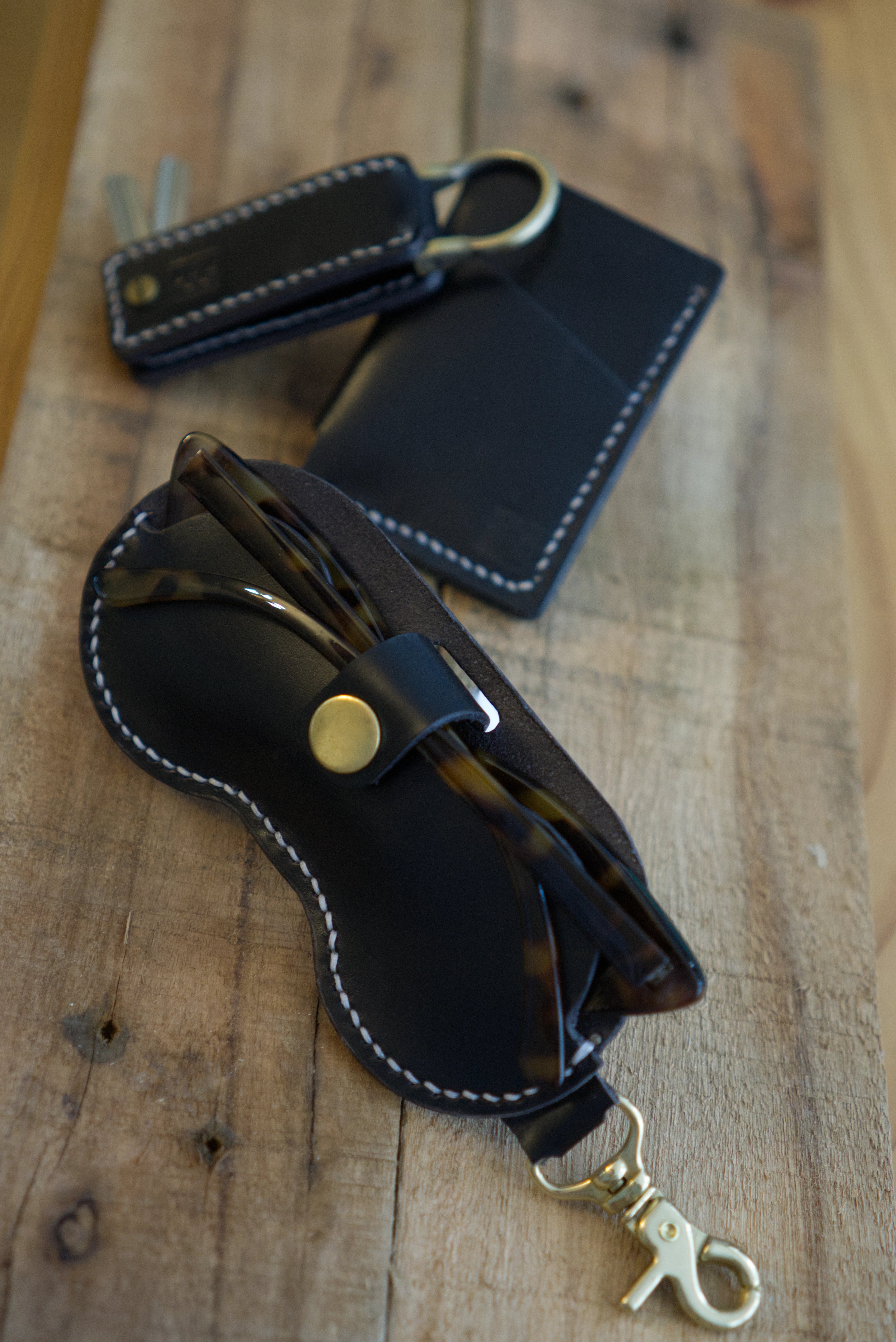 Black leather glasses case with brass strap on a wooden surface. Key holder & wallet in background