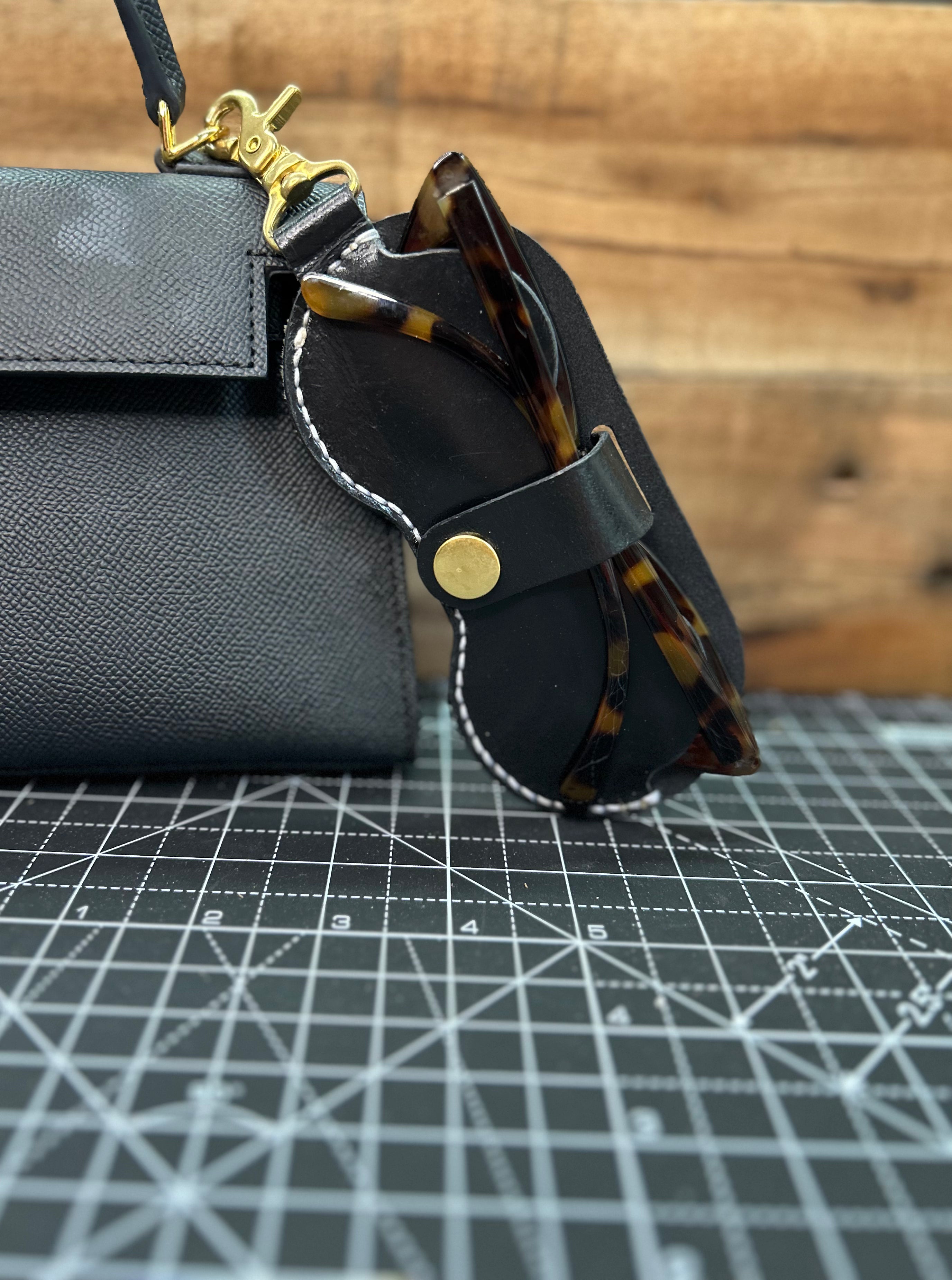 Black leather glasses case with tortoiseshell glasses on a grid surface.