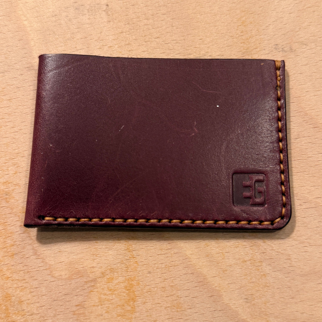 A dark reddish-brown leather bifold wallet with contrast stitching and an embossed 'EG' logo in the bottom right corner, resting on a light wood surface.