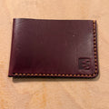 A dark reddish-brown leather bifold wallet with contrast stitching and an embossed 'EG' logo in the bottom right corner, resting on a light wood surface.