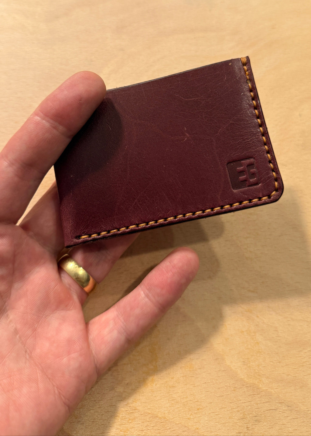 A hand holding a dark burgundy leather card holder wallet with contrast stitching and a small embossed logo on the bottom right, set against a light wood background.