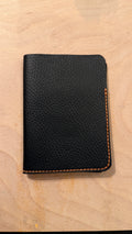 A black textured leather passport cover with orange contrast stitching, shown closed on a light wood background.