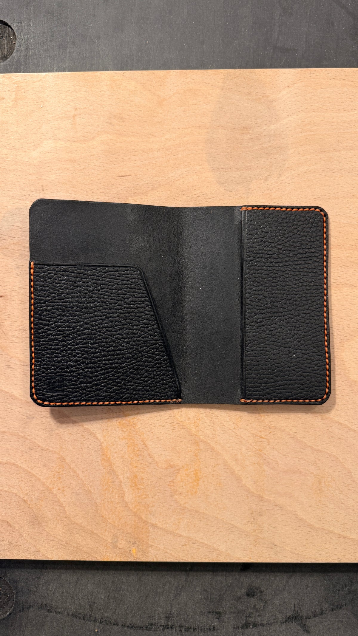 A black textured leather passport  wallet with orange contrast stitching, shown open on a light wood surface.