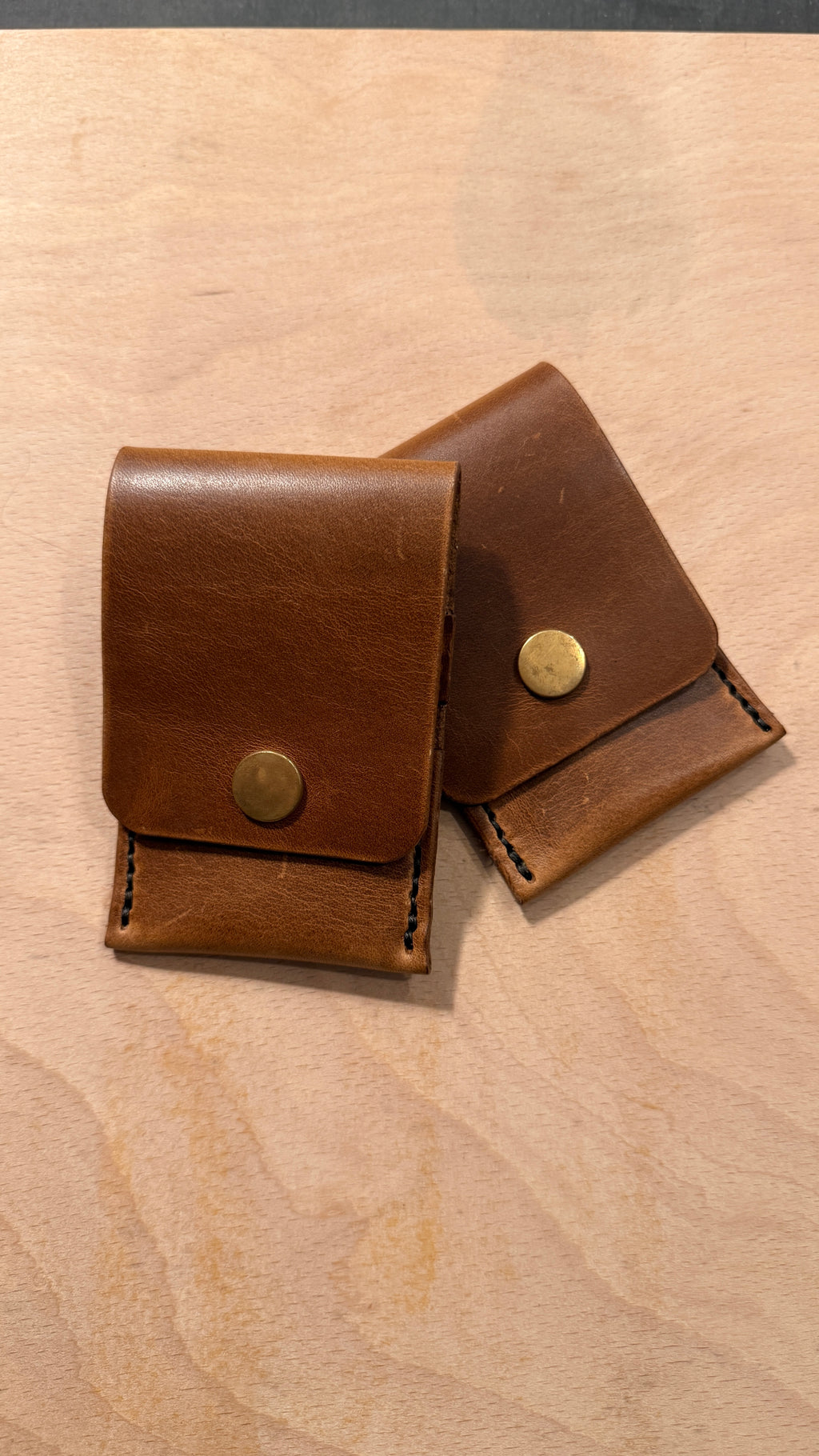 Two small brown leather card cases with brass snap closures, arranged on a light wood surface.