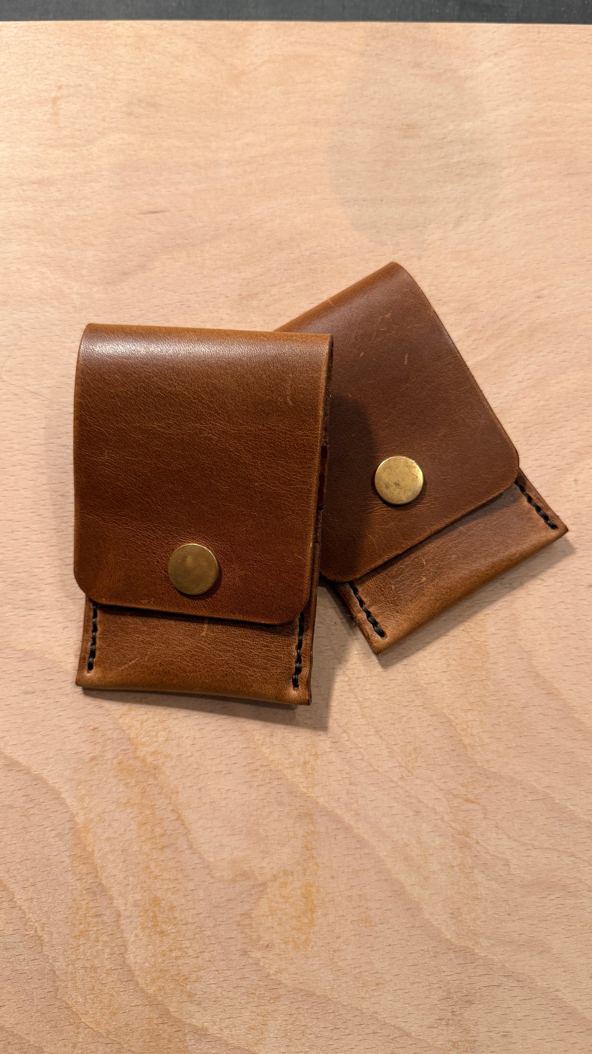 Two small brown leather card cases with brass snap closures, arranged on a light wood surface.