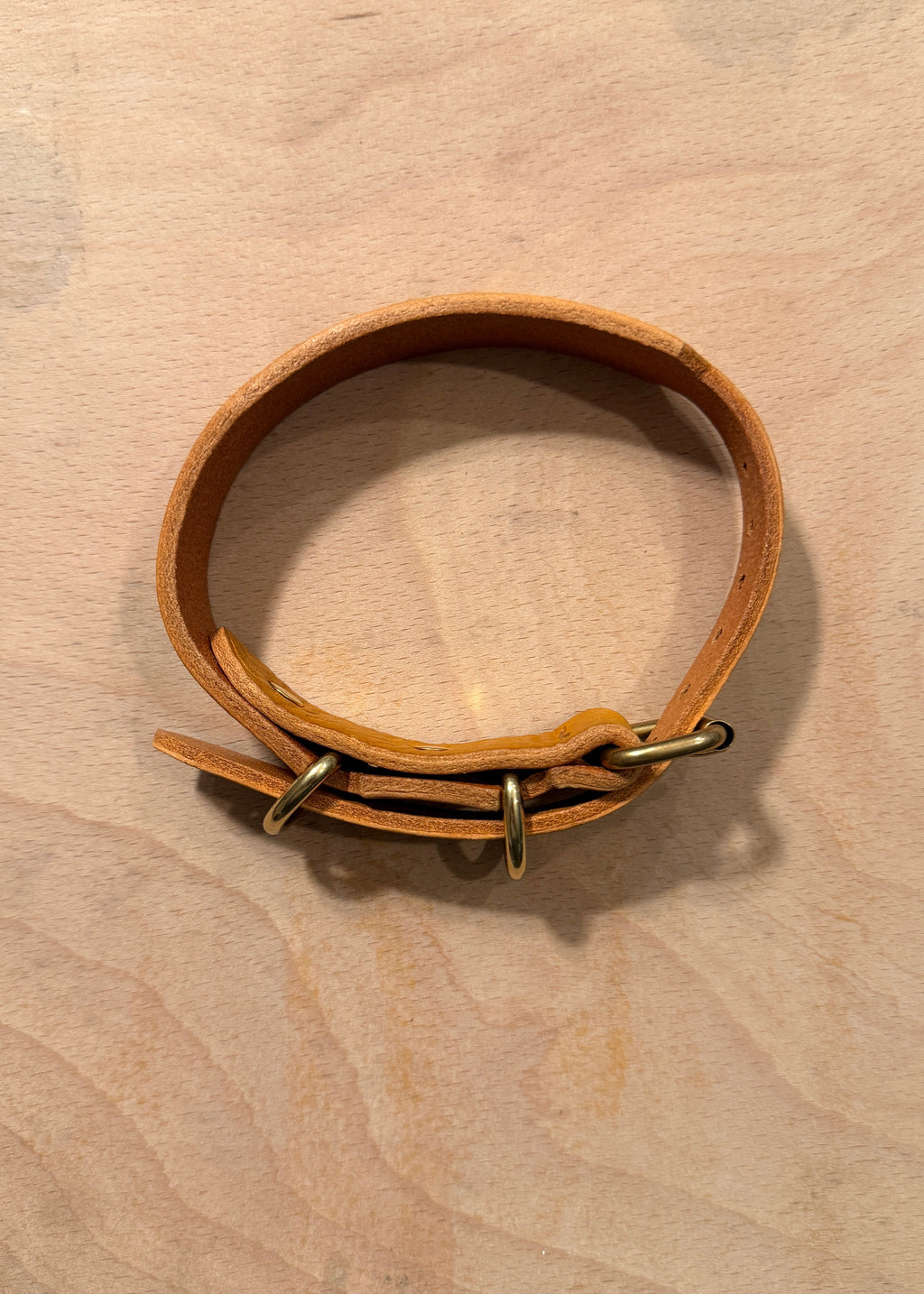 A light brown leather dog collar with brass hardware, displayed on a light wood background.