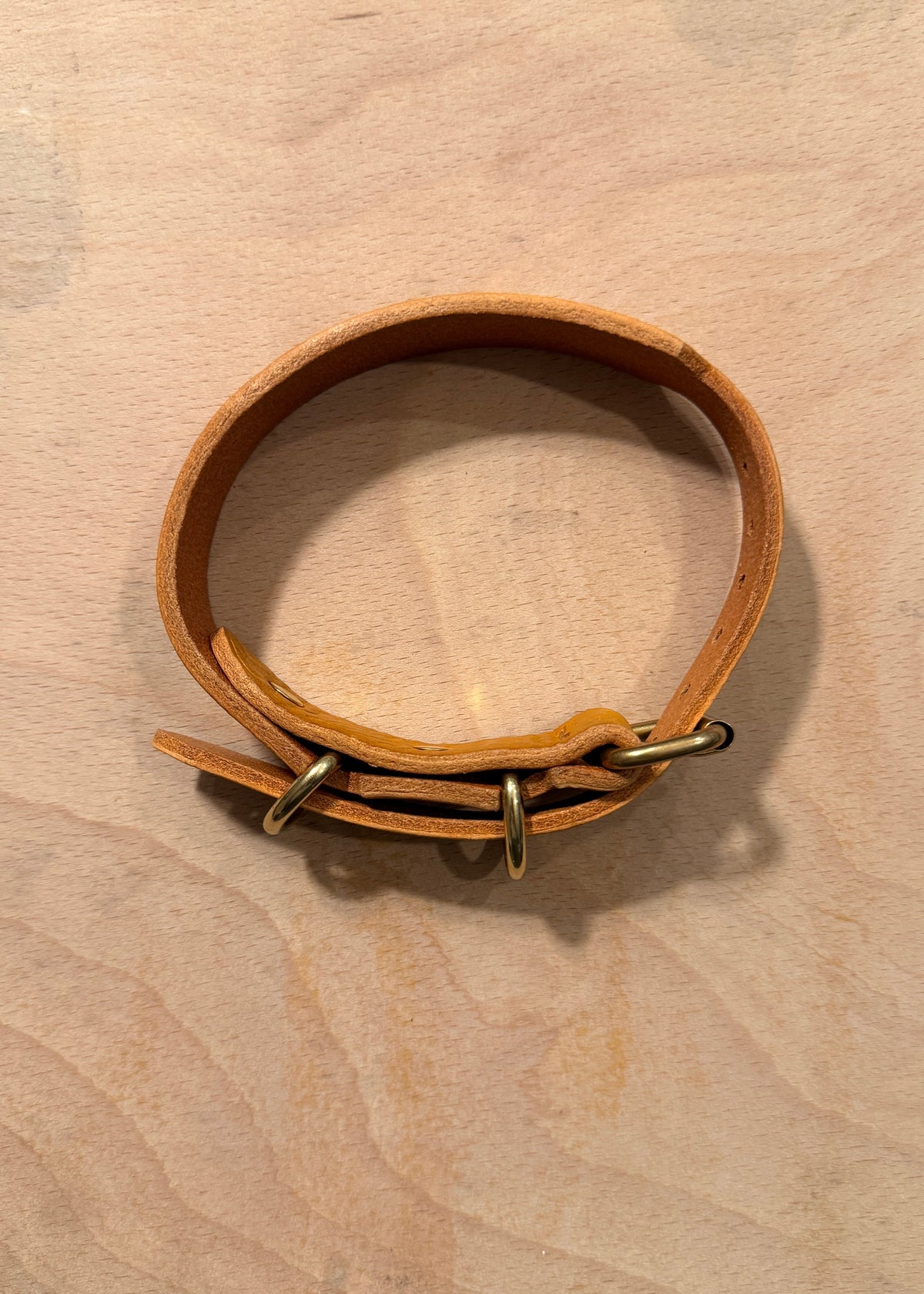 A light brown leather dog collar with brass hardware, displayed on a light wood background.