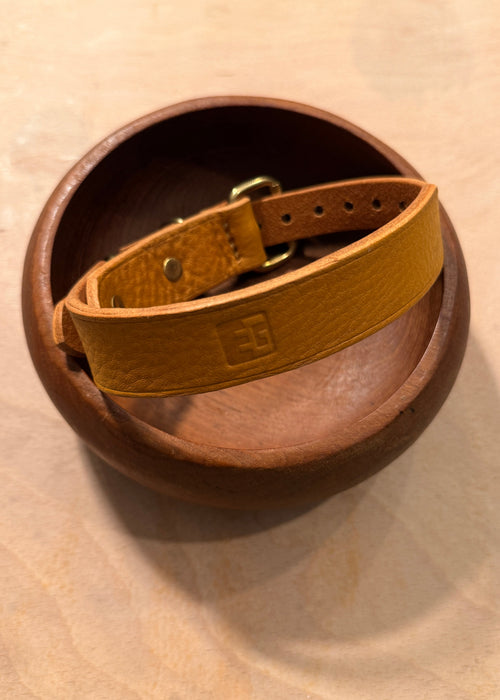A tan leather dog collar with a brass buckle, resting inside a round wooden bowl on a light wood surface.