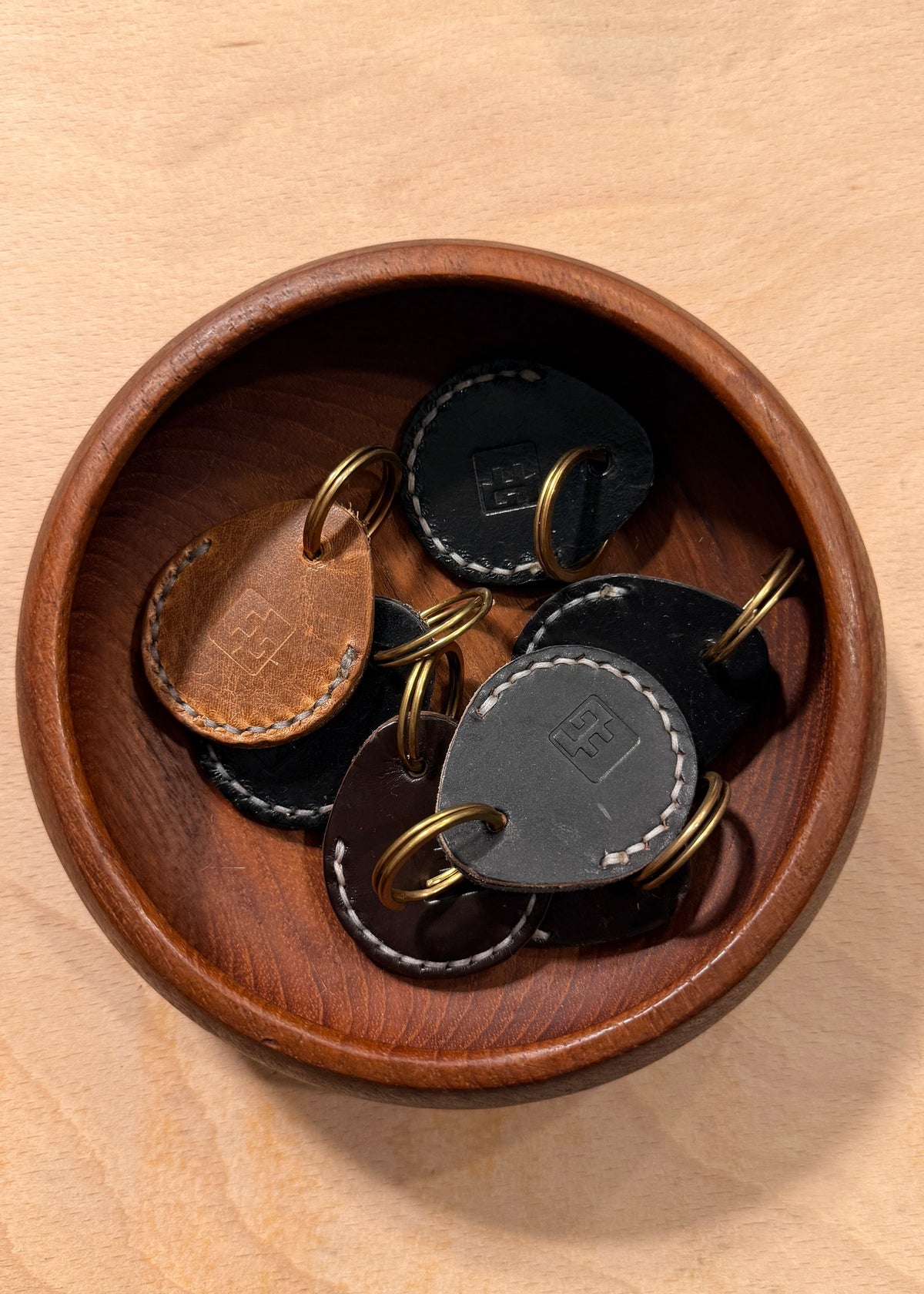 Several round leather key tags with brass key rings resting inside a round wooden bowl on a light wood surface.