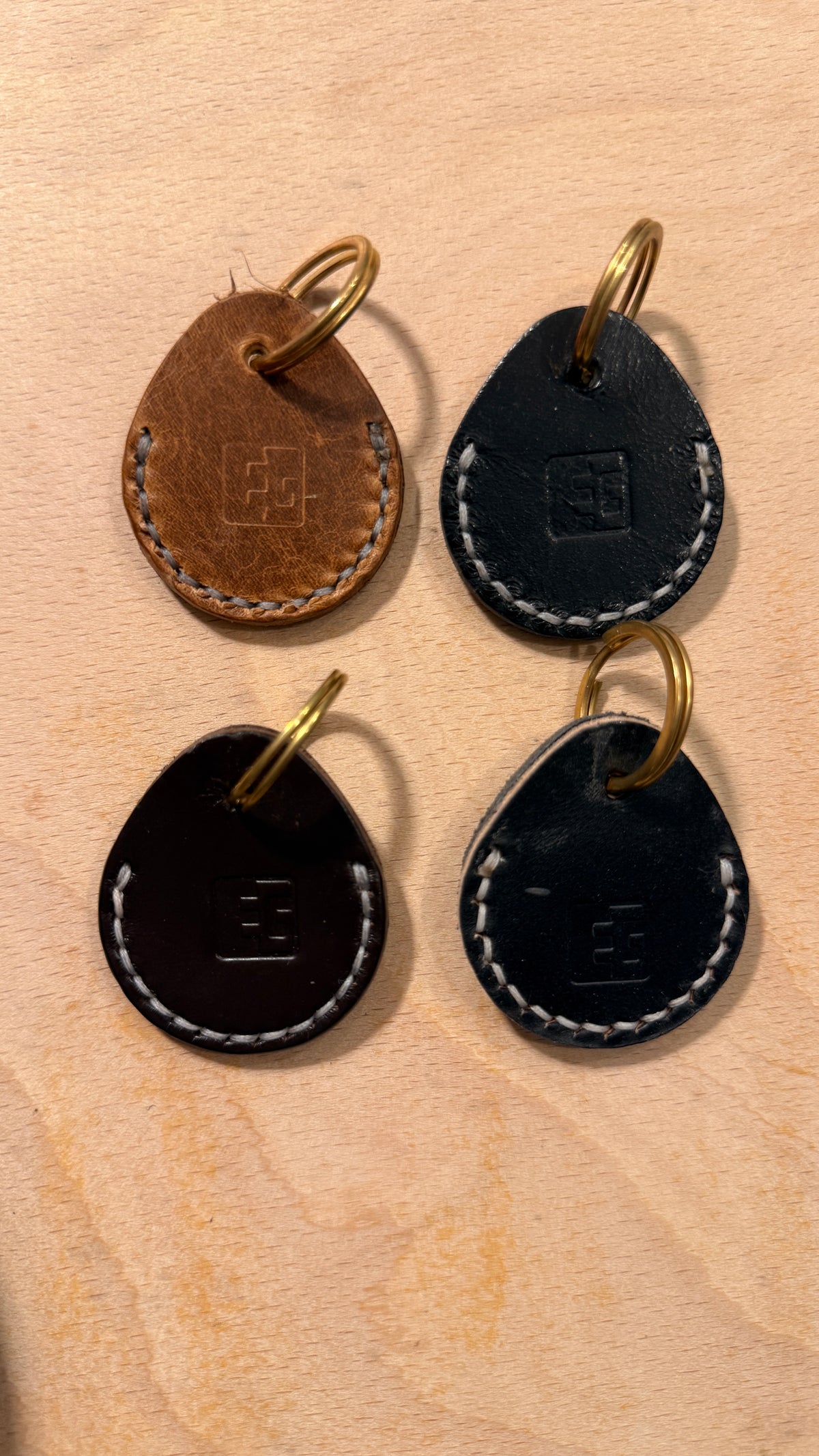 Four teardrop-shaped leather keychains arranged in a 2x2 grid on a light wood background. The top-left keychain is brown with gold hardware, and the other three are black with white contrast stitching and gold hardware.