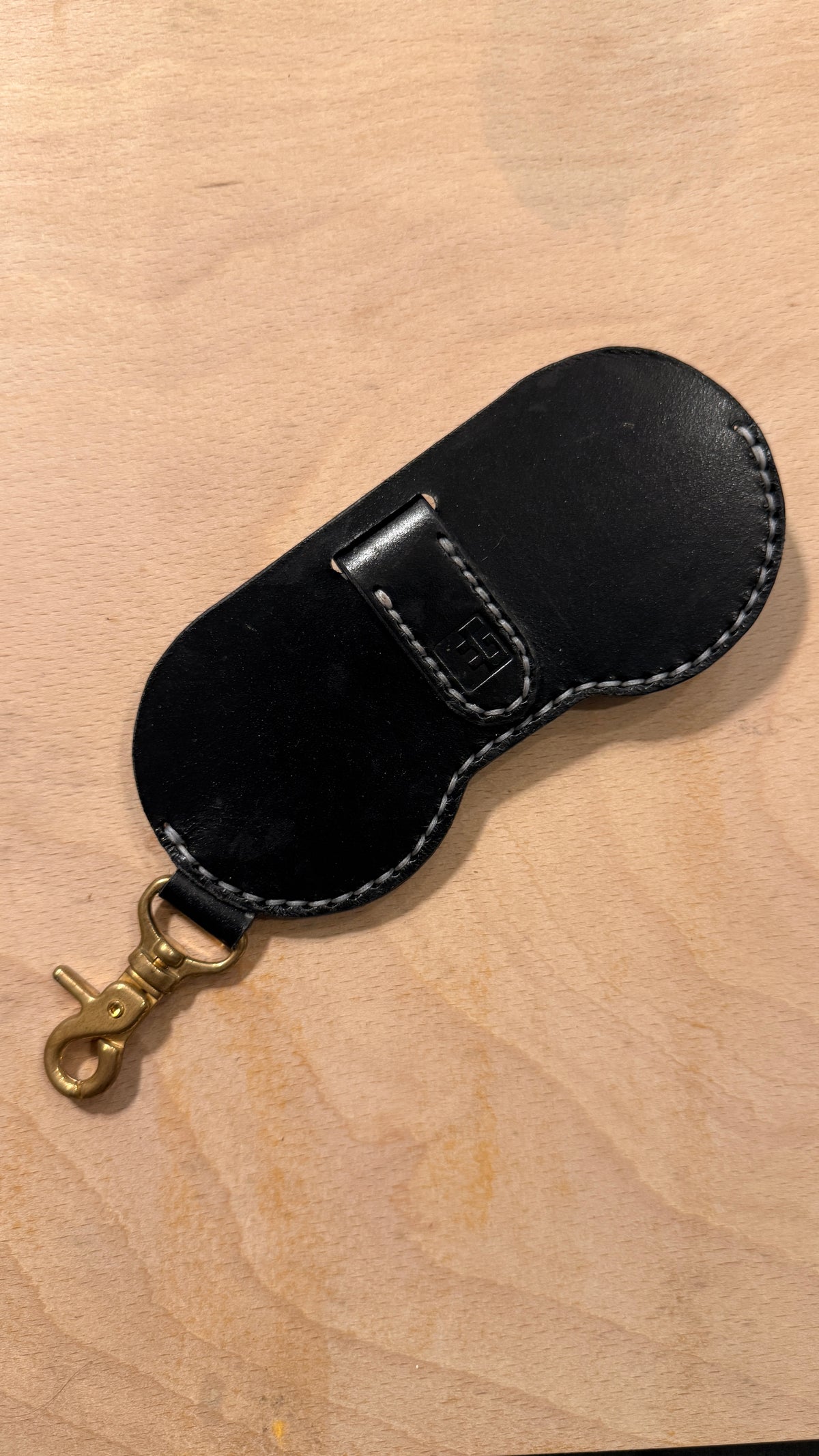 Black leather Holstein glasses case with white saddle stitching, embossed Everbound logo and a solid brass lobster claw clasp, laid flat on a wooden surface