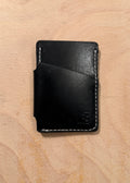 A minimalist black leather card holder wallet with white contrast stitching, shown closed on a light wood grain background.
