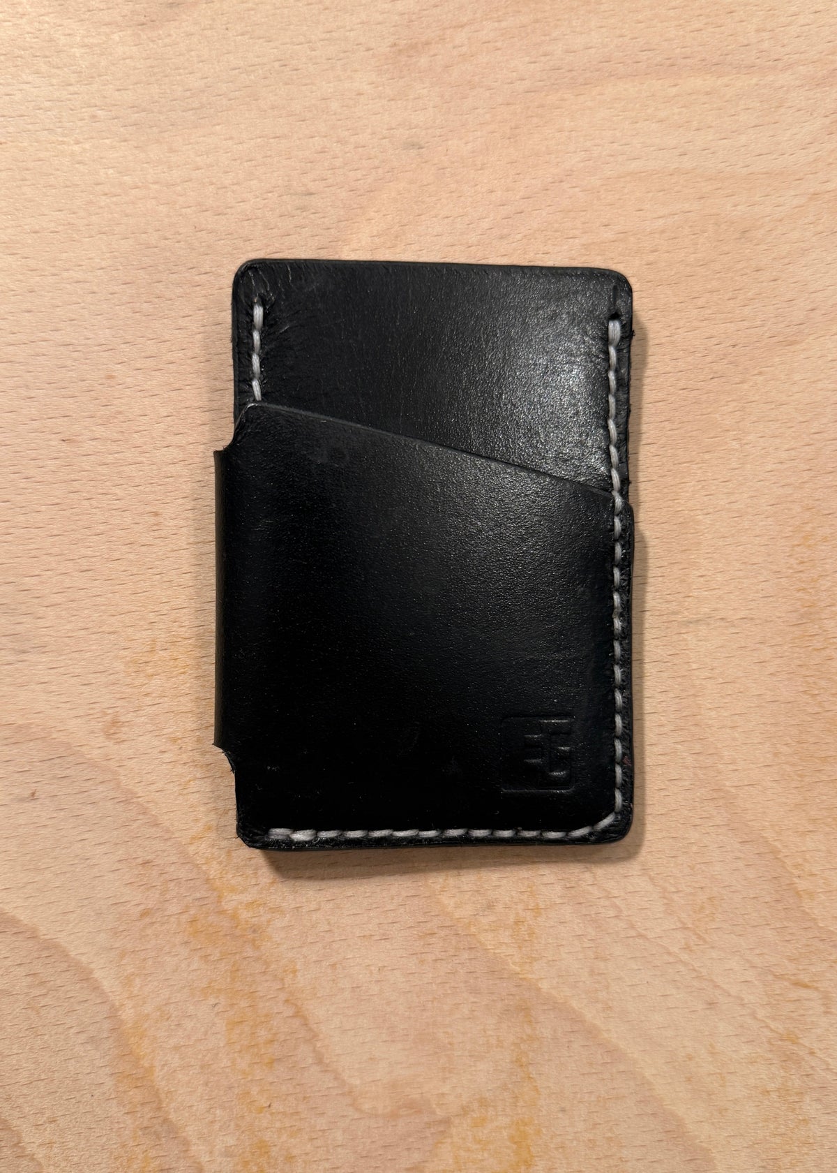 A minimalist black leather card holder wallet with white contrast stitching, shown closed on a light wood grain background.