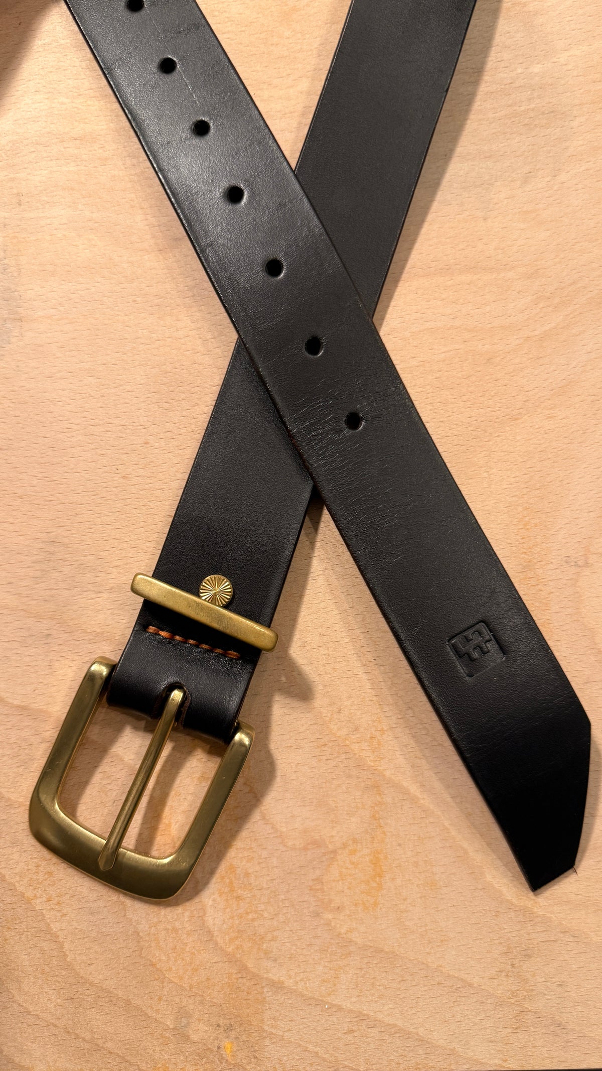 A black leather belt with a brass buckle, laid out in an X-shape on a light wood background.