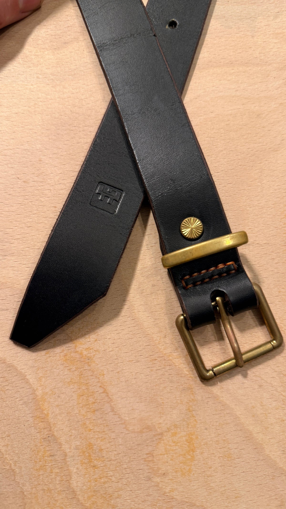 A close-up of a black leather belt with a brass buckle and a small gold-toned snap detail, laid on a light wood surface.