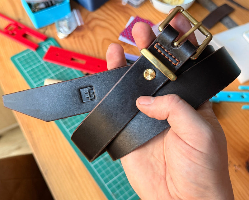 Hand holding a black leather belt with a brass buckle on a wooden table.